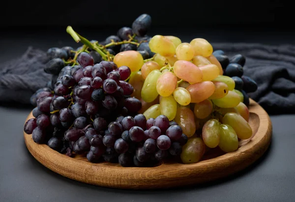 Black and green grapes in a wooden round plate on a black table, top view