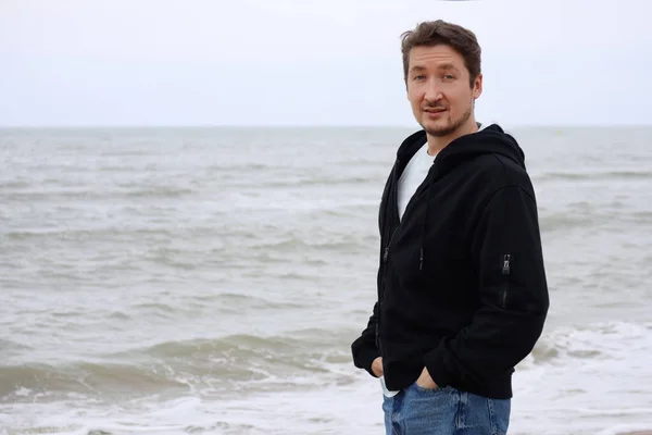 A young man in a black hoodie on the beach against the backdrop of the sea. Brown hair, blue eyes, smiling