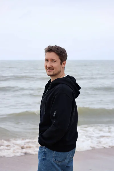 A young man in a black hoodie on the beach against the backdrop of the sea. Brown hair, blue eyes, smiling