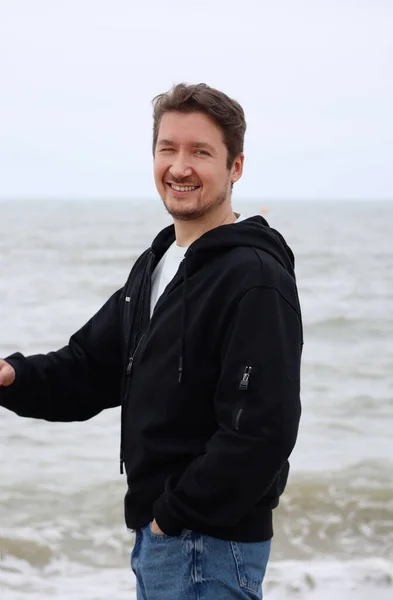 A young man in a black hoodie on the beach against the backdrop of the sea. Brown hair, blue eyes, smiling