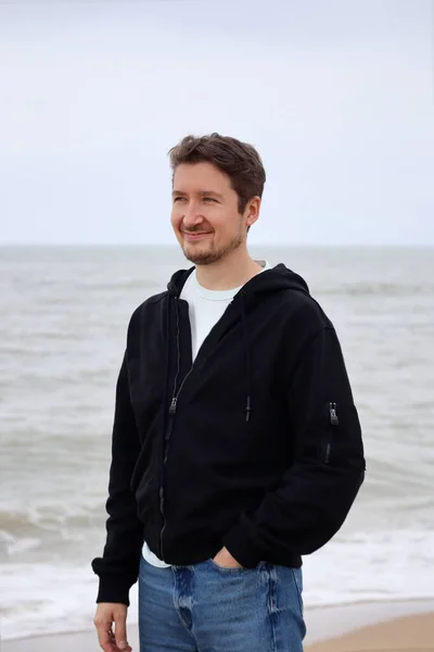 A young man in a black hoodie on the beach against the backdrop of the sea. Brown hair, blue eyes, smiling