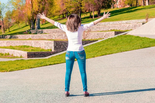 Arkana bak kot pantolon ve beyaz bluzlu genç kadın ellerini kaldırdı ve parkta duruyor.