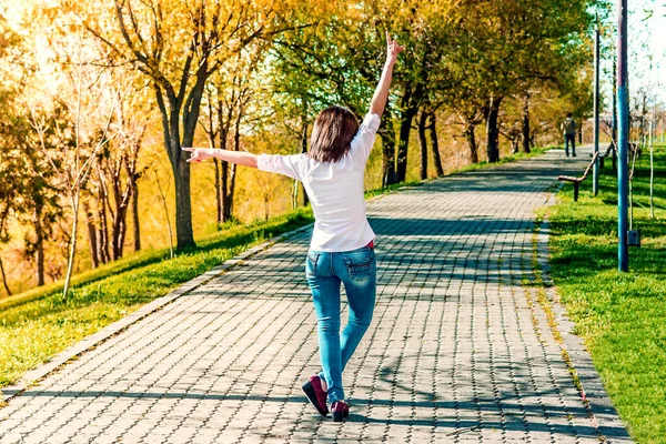 Arkana bak kot pantolon ve beyaz bluzlu genç kadın ellerini kaldırdı ve parkta duruyor.