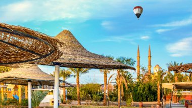 Güneşli bir plajda saman şemsiyesi ve Mısır bayrağı renginde balon, arka plandaki El Sahaba camisinin minareleri üzerinde dalgalanıyor.