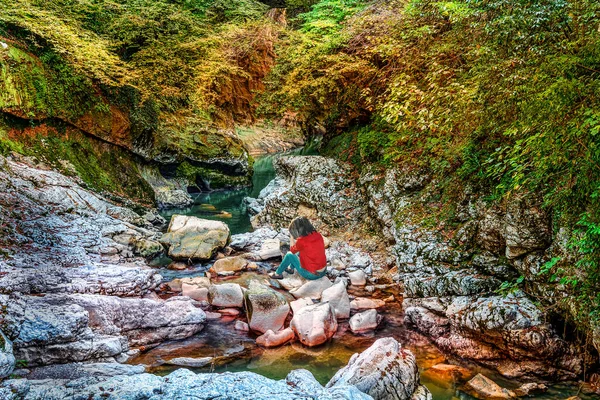 Young sad girl in jeans and a red blouse is sitting on the rocks on a mountain river with her head down.