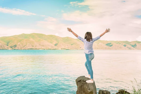 A young girl in a blue blouse is standing on the stone shore of the lake spreading her arms