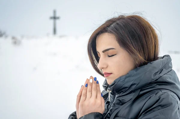 A beautiful young woman prays with her eyes closed, her hands folded with her palms, a girl against the background of a Christian cross set on a hill. Winter season