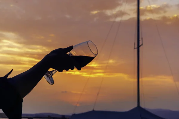 Silhouette of a female hand holding a glass of red wine in her hand on the seashore against the backdrop of a beautiful evening sunset