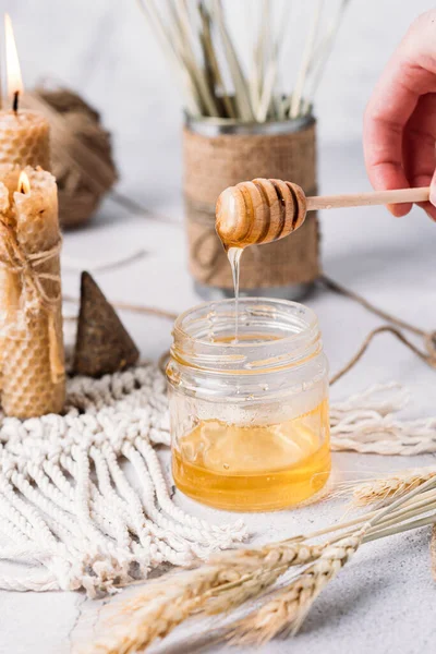 Liquid transparent honey in a glass jar hand holding a honey stick honey dripping wheat macrame concrete background