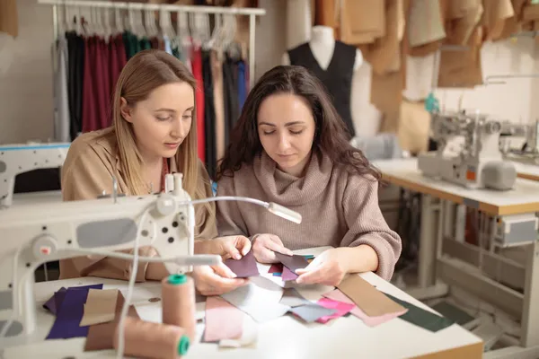 Zwei junge Schneiderinnen oder Designerkolleginnen arbeiten und diskutieren über Modeprojektgestalter und betrachten Stoffmuster für Kleidung, Beruf und Beruf — Stockfoto