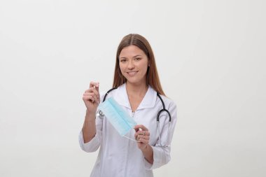 Üniformalı genç bir kadın doktor beyaz arka planda tıbbi bir maske tutuyor. Hemşire başparmağını gösteriyor..