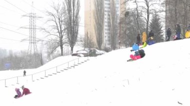 Ukrayna, Kyiv - 30 Ocak 2021: İnsanlar çok katlı bir binanın avlusunda kar tepesinde kayıyorlar. Kışın karlı bir günde plastik bir kızakla kayan çocuklar. Mutlu bir çocukluk. Dışarıda kar yağıyor. Tatil çocukları eğleniyor
