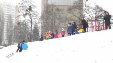 Ukrayna, Kyiv - 30 Ocak 2021: İnsanlar çok katlı bir binanın avlusunda kar tepesinde kayıyorlar. Kışın karlı bir günde plastik bir kızakla kayan çocuklar. Mutlu bir çocukluk. Dışarıda kar yağıyor. Tatil çocukları eğleniyor