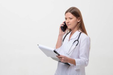 Tıp önlüğü içinde telefonla konuşan genç bir kadın doktor ve beyaz arka planda steteskop ve dosya klasörü.