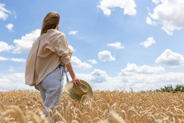 Yazın buğday tarlasında poz veren bir kadın. Elinde altın buğday tarlasının arka planında şapkasını tutan bir kadın. Arkadan manzara..