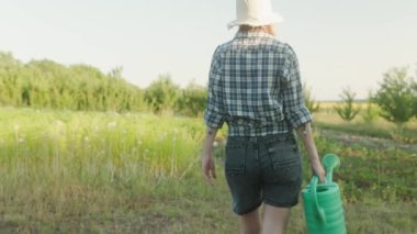 woman farmer walkinfg inspects the gatden. Girl holds watering can in hands for plants. Casual summer rural clothes denim clothes. Agricultural concept. Back view