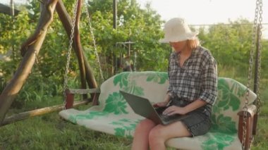 Young adult woman working outdoors in her garden with laptop. Freelancer using laptop computer while sitting in cozy garden swing chair at backyard of her house