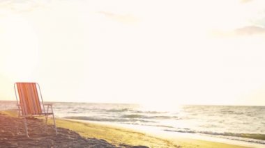 Güneşin batışında (gün doğumunda) bir güverte sandalyesi boş bir kumlu sahilde durur. Güneş, suya yansıyor. Sabit Kamera