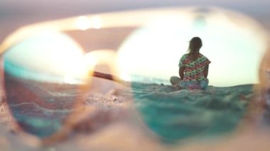 Şafak vakti kumlu bir plajda oturan bir kız (güneş gözlüklerinin yansımasında). Sabit Kamera