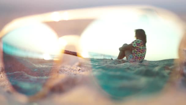 女の子は夜明け に砂浜に座っています サングラスの反射で見る 固定カメラ — ストック動画