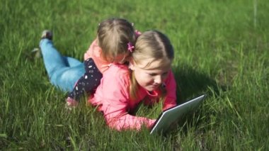Bahar. Güneşli bir hava. Yeşil çimenli tepe. İki kız elinde tabletle çimlere uzanıyor. Sabit Kamera