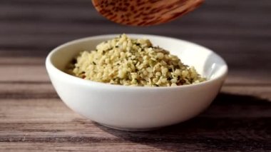 Organik kenevir tohumları küçük beyaz bir tabağa dökülüyor ve tahta bir arka planda kaşık var.