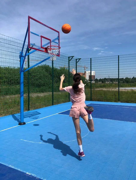 A young woman with a basketball on an outdoor blue basketball court. Photo in motion, girl is throwing a basketball into the hoop