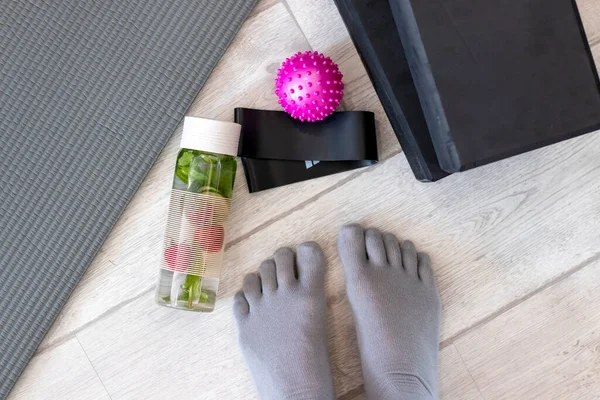 A girls feet in toe socks got ready to exercise on a mat with tape, a ball, yoga blocks and cucumber water