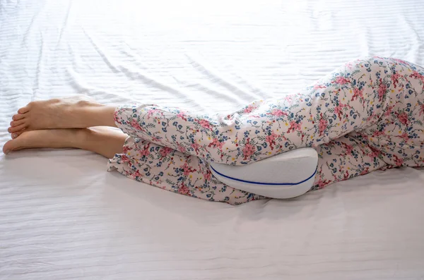 Woman in pajama pants with an anatomical pillow between her legs and knees, lying on a bed with white sheets