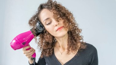 Genç bir kadın saç kurutma makinesiyle saç maşası yapıyor. Saç bakım konsepti. Bir kız dalgalı saçlarını şekillendiriyor