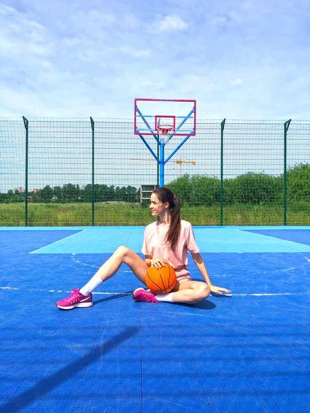 Slim young woman sitting on a blue field with a basketball. Vertical photo.