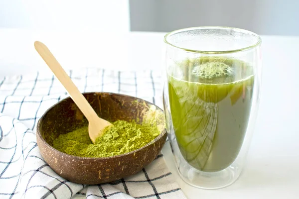 Matcha green tea in a double-bottomed glass with a coconut bowl filled with matcha powder on a white background