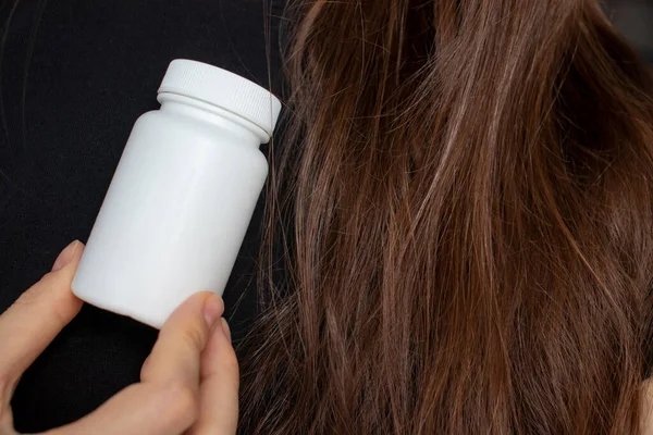 A woman holds an empty bottle of supplements and vitamins in her hand against a background of healthy hair and nails. The concept of healthy hair care
