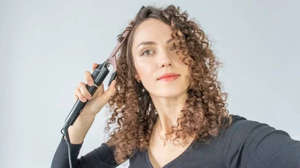 Curly hair. Beautiful brunette woman with curly hair, curls her hair, uses curling tongs to get fine curls. Hairstyles and hairdressing tools