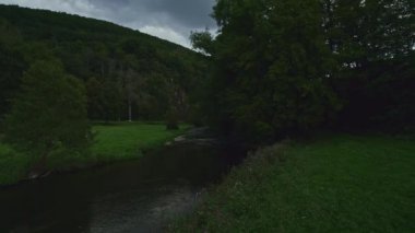 Low view over the peaceful calm river Sauer and low underneath the trees at dusk, meadows with fresh green grass, forests surrounding the meadow and a blue sky in the background, in the Luxembourg forest. Drone aerial view.