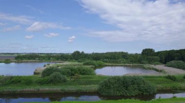 İnsansız hava aracı bir çayırın üzerinden uçar ve hendek kazar. Ağaçlar, çalılar ve çalılarla dolu bataklık bir arazide göletin üzerinde ve bisikletçilerin üzerinde uçmak. Hollanda manzarası üzerinde mavi gökyüzü ve beyaz bulutlar 4K