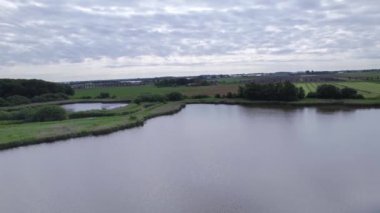 Çiftlik arazisi ve doğası olan kırsal bir kırsal bölge. İnsansız hava aracı su ve çayırlar üzerinde uçuyor. Gölet sazlıklar, bir park ve bir orman ile çevrilidir. Bulutlu mavi gökyüzü. Tarım sektörü manzarası 4K