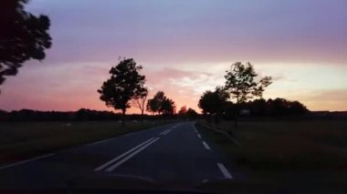 Gece geç saatlerde polis korkuluklarıyla, gün batımından sonra arabalarla eyalet yolunda giden POV arabası. Kırsal alandaki tarlaları ve tarlaları geçmek. Turuncu, kırmızı ve mavi gökyüzü güzel renkli. Bakış açısı 4K