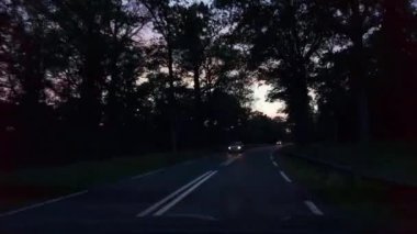 Gece geç saatlerde, gün batımından sonra bariyerlerle eyalet yolunda giden POV arabası. Kırsal bir arazide tarlaları ve tarlaları geziyorlar. Turuncu, kırmızı ve mavi ile güzel renkli gökyüzü