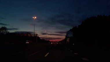 POV gece geç saatlerde otoyolda arabalarla geziyor. Gün batımından sonra yol boyunca bariyerler, bariyerler ve sokak lambalarıyla. Güzel renkli turuncu, kırmızı ve mavi gökyüzü. Torpidodaki kamera. Bakış açısı 4K