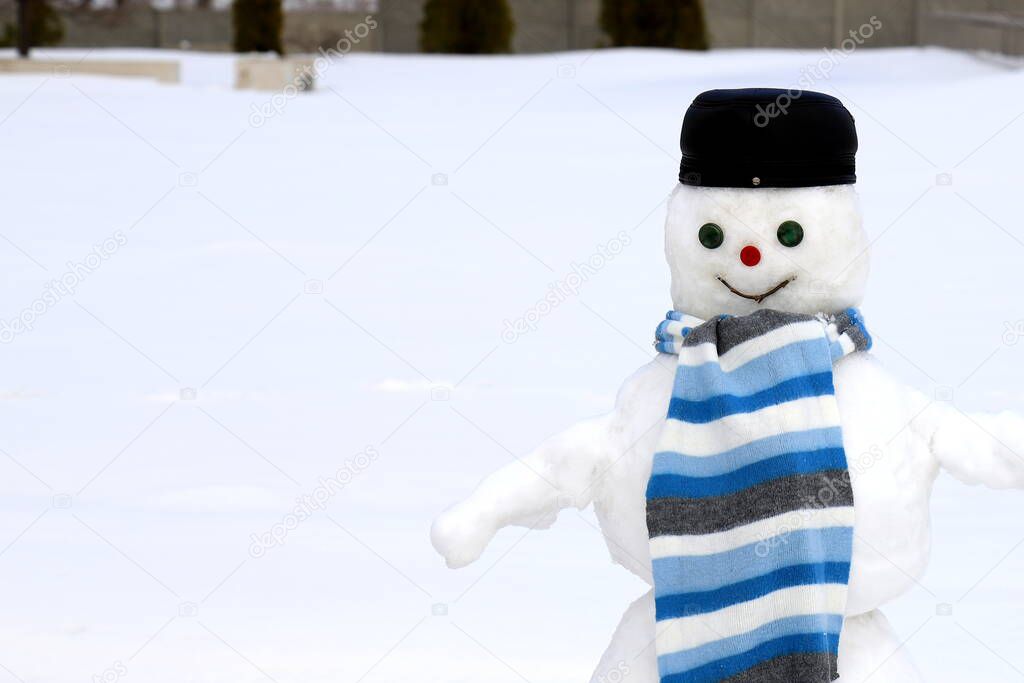 A funny snowman stands on snow covered playground in winter among white snow. A snowman in black cap and a blue scarf, Christmas and New Year holidays, children entertainment and sports,