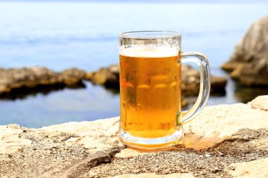 Glass of light beer near sea, close-up. Misted mug, pint of cold beer stands against background of blurred sea landscape in cafe, restaurant, bar, pub, Herceg Novi, Montenegro