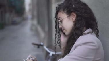 Long Curly-Haired Moroccan Girl Looking On Her Wristwatch While Talking On A Smartphone. Zoom out - Horizontal