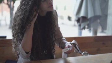 Beautiful Young Woman In Eyeglasses Puts On Bluetooth Earbuds While Sitting Outdoor. medium shot - Horizontal