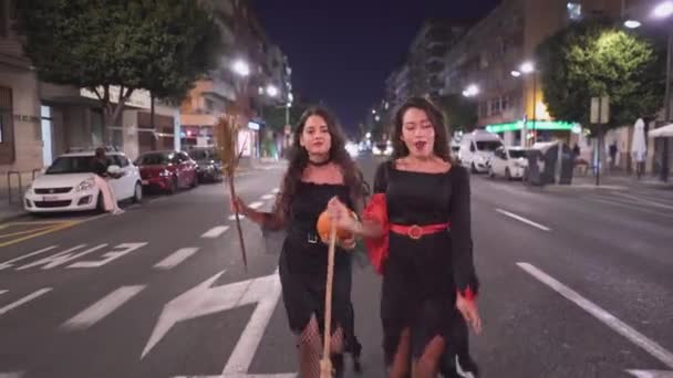 ブロムスティック付きの魔女の衣装を着た2人の素敵なコロンビア人の女の子 1人はカボチャを保持しています夜 バレンシア スペインの通りを歩くのを楽しんでいます フルショット 横4K — ストック動画