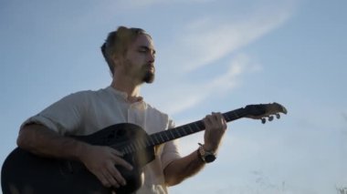 Açık havada gitar çalan İspanyol sakallı adam güneş ışığıyla aydınlatılmış mavi gökyüzüne karşı.