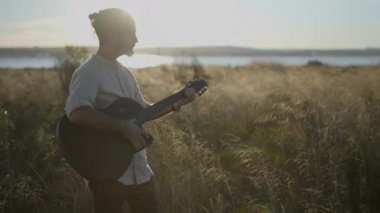 İspanyol erkek gitarist Sihouette gün batımında göl kenarında çimenli bir çayırda akor çalıyor ve şarkı söylüyor.