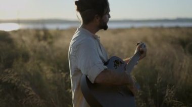 Man Guitarist İspanya 'da çimenlik alanda duruyor ve siyah akustik gitarla güzel güneş ışığına karşı akorlar çalıyor - yavaş çekim profil görüntüsü - yatay 4K video