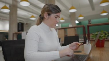 Ciddi anlamda başarılı genç bir kadın çalışma alanında oturuyor, elinde bir kredi kartı tutuyor ya da pin kodunu giriyor, dikkatle dizüstü bilgisayar ekranına bakıyor, internetten sipariş veriyor. -
