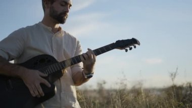 Sakallı İspanyol gitaristin portresi, sonbaharda gün batımında açık gökyüzünün altında gitar çalıyor.
